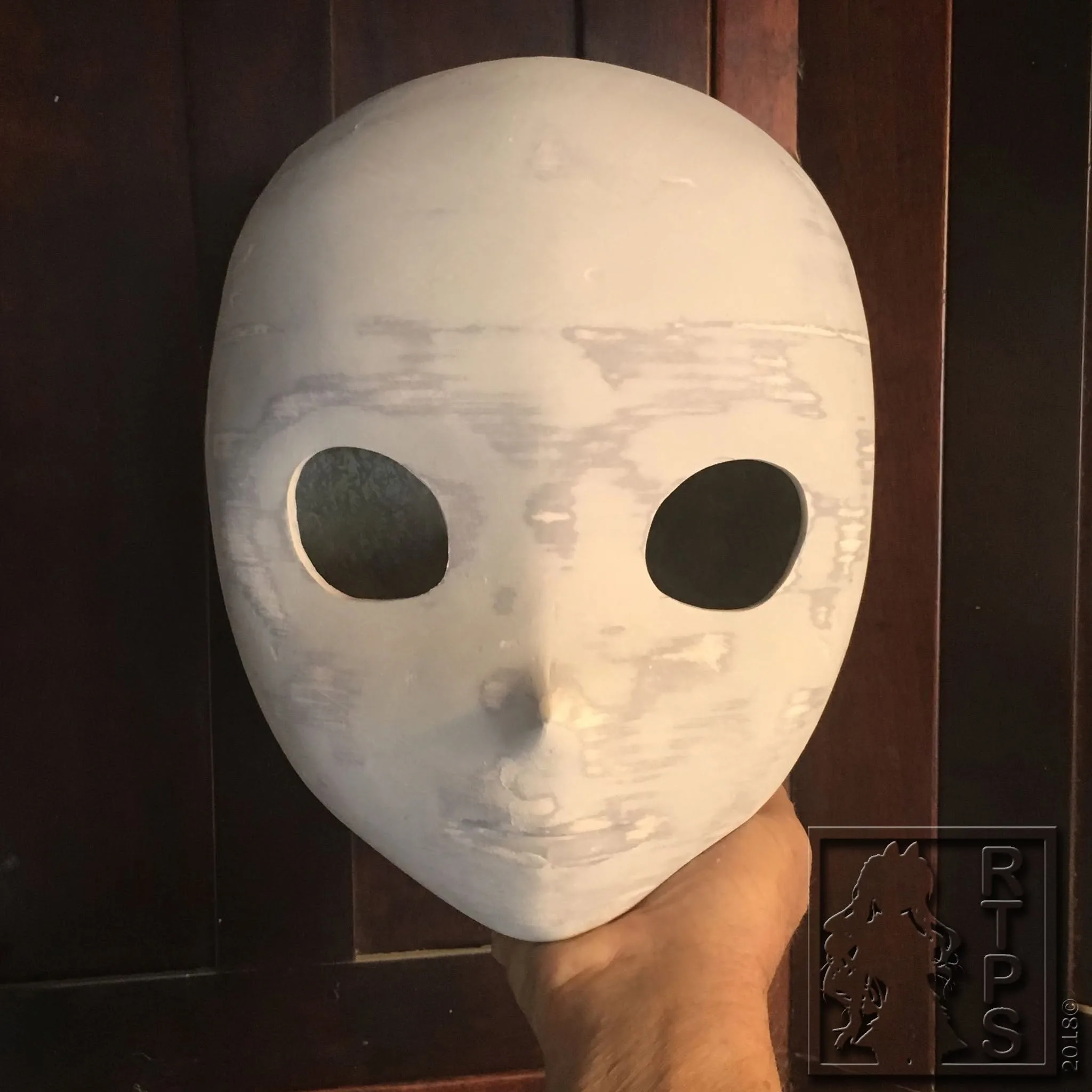 Unfinished sanded Ride the Pig Studio mask shell held against a wooden door, 2018